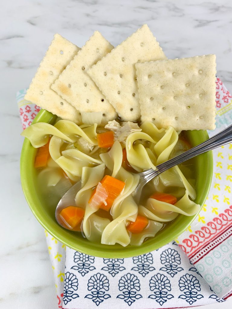 On a white marble table, there's a napkin with colorful designs. On top of the napkin is a green bowl with chicken noodle soup. There's a spoonin the bowl and crackers on top of the bowl.