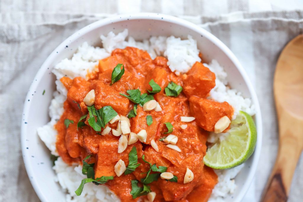 Sweet potato curry in a white bowl over rice. with a wooden spoon on the right. how to make red sweet potato curry