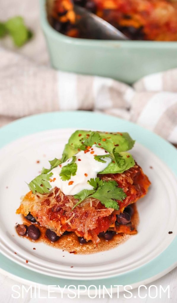 a piece of sweet potato and black bean casserole on a green and white plate garnished with avocado and red pepper flakes and parsley.