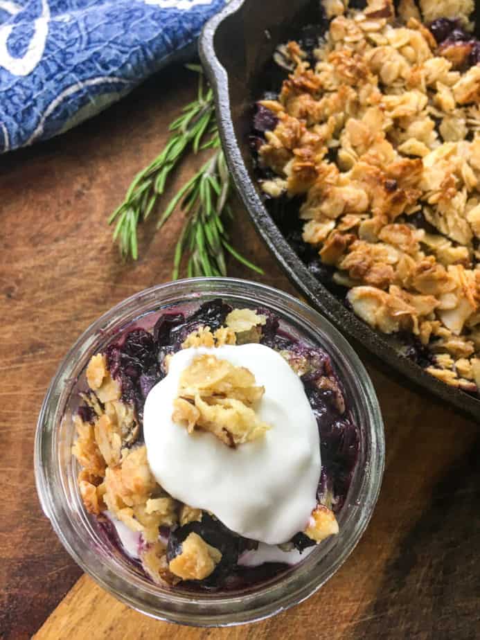 Blueberry crisp in a small mason jar with whipped cream and granola on top. The jar sits on a wooden table, next to a cast iron pan with more crisp in it. 