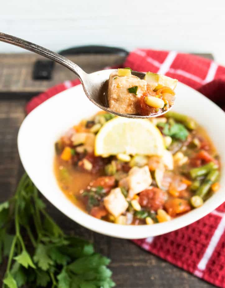 On a wooden table sits a red napkin with a waffle texture and white lines on it. On that sits a white bowl filled with fish stew, topped with a lemon slice. Next to the bowl are some fresh herbs. There is a spoon above the bowl with a bit of the fish stew in it. 