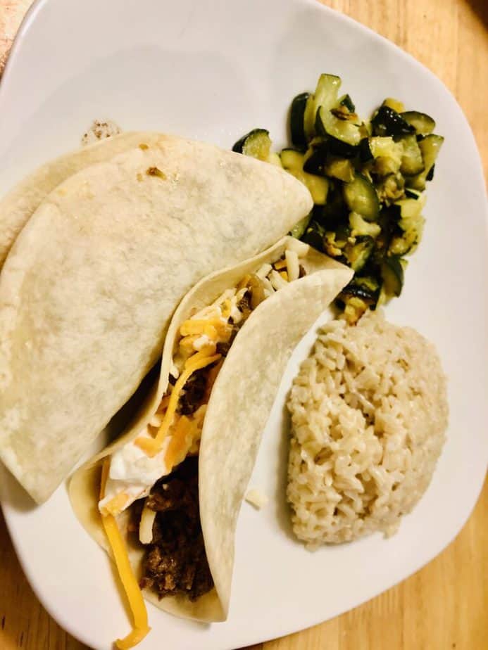 flour tortilla shell with steak, sour cream and cheddar cheese on a white plate with a side of brown rice and zucchini