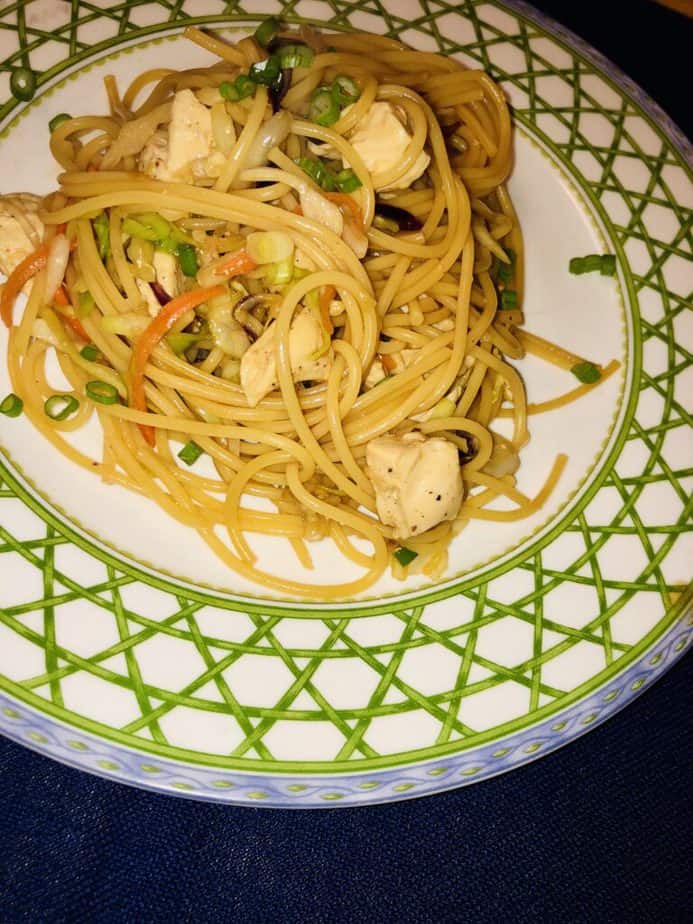 Blue background. White plate with a green lattice rim and a blue edge. On the plate is pasts with chicken, cabbage and scallions. 