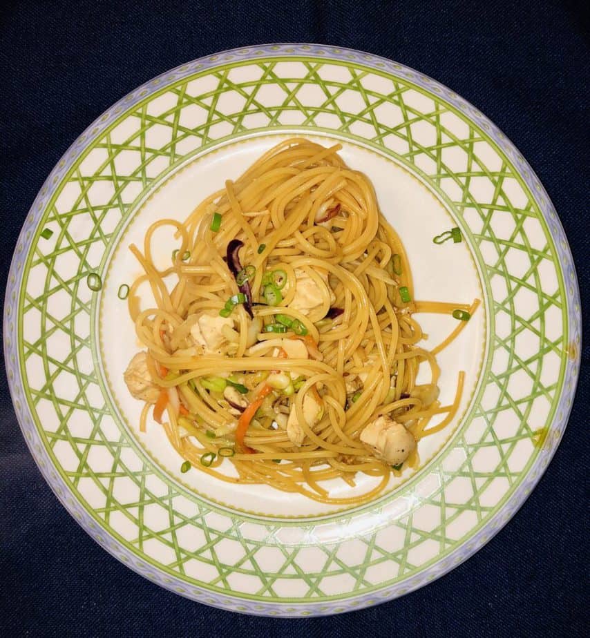 Blue background. White plate with a green lattice rim and a blue edge. On the plate is pasts with chicken, cabbage and scallions. 