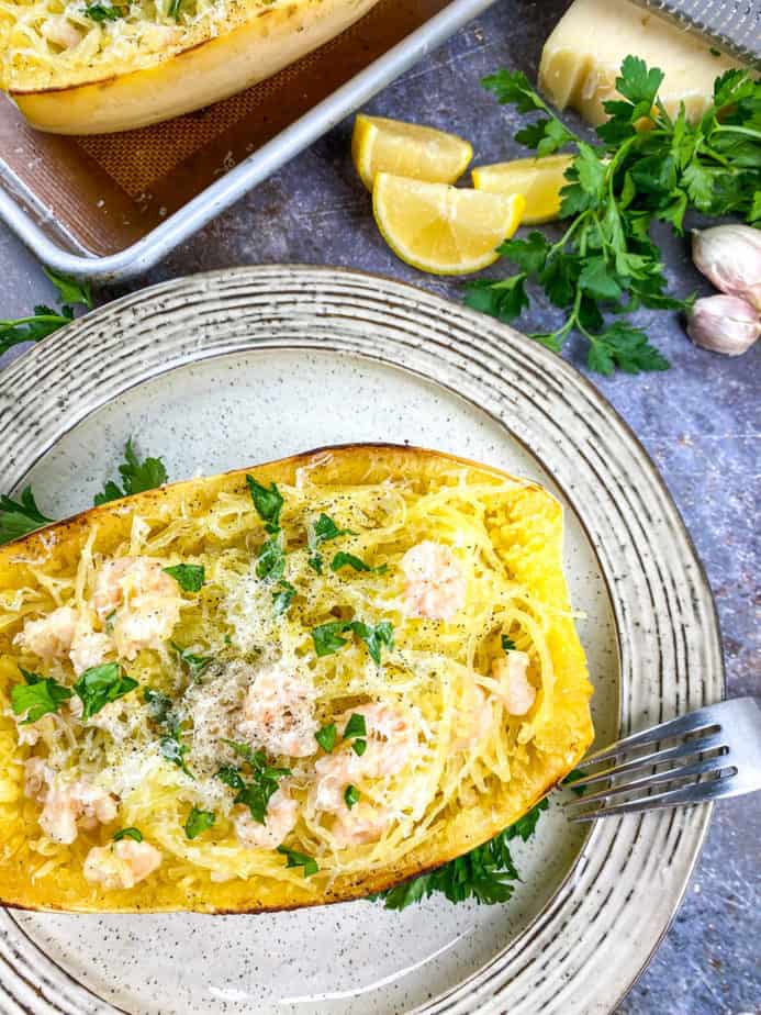 A white plate with brown speckles and brown lines on the rim. On the plate is half of a spaghetti squash with shrimp and parsley on top.  In the background are cut up lemons and sprigs of whole parsley. 