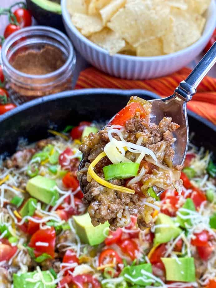 A cast-iron skillet with beef on the bottom and covered with cherry tomatoes, chunks of avocado, and a sprinkling of cheddar cheese. On the top left there are cherry tomatoes on the vine. Next to that is a small glass bowl with a spoon in it and some spices. To the right of that is a white bowl with corn chips in it. Everything is on a wooden table. There's a red napkin with orange and yellow thin stripes on it to the right of the skillet. There's a spoonful of the taco meat as the focal point of the picture. 