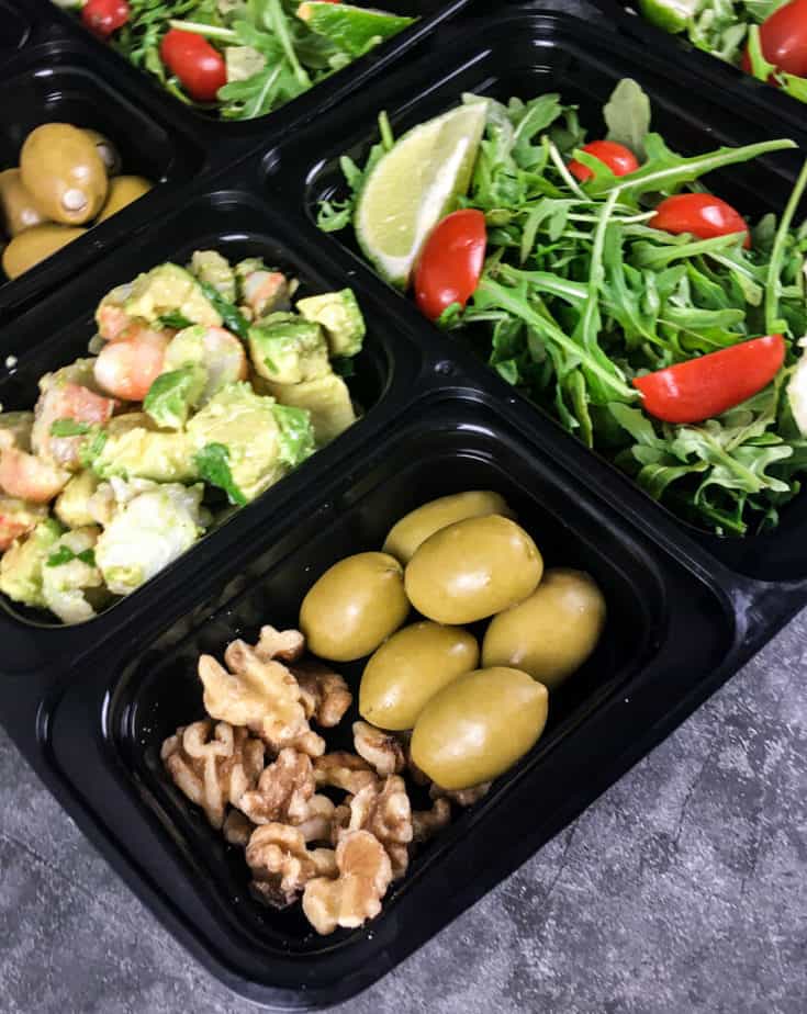 A bento box with field greens, sliced grape tomatoes and a lime wedge in one square, the keto shrimp avocado in one square, green olives and walnuts in a square, green olives in another square