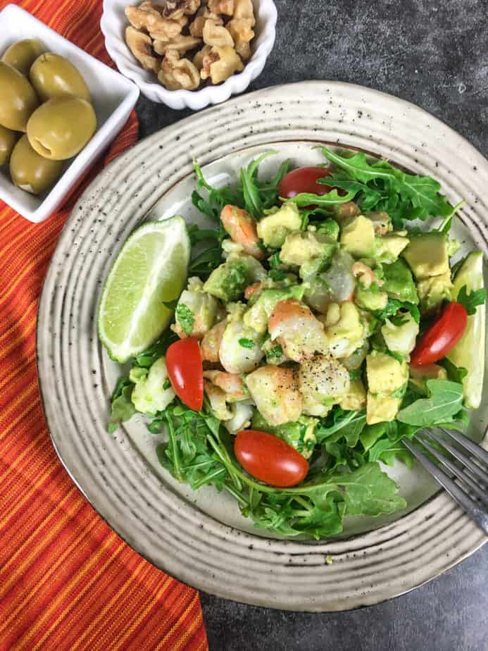 White plate on a grey marble table with a red napkin with red, yellow and orange stripes. On the plate is a bed of field greens. The keto shrimp avocado salad is on that, with a wedge of lime. There's an upside fork on the edge of the plate. There's a small, square bowl on the top left with green olives in it. 