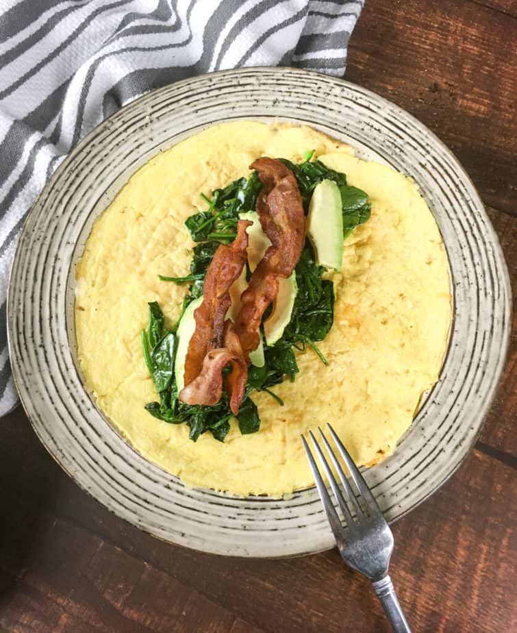 An off-white plate sits on a wooden table. There's a white napkin with grey stripes on the upper left, under part of the plate. On the plate is an open-faced egg wrap. In the middle is a pile of spinach, topped with avocado and then bacon. There's an upside down fork on the edge of the plate. 