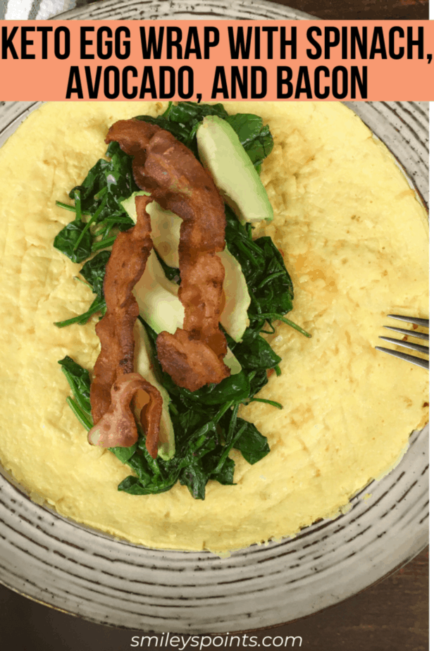 On the plate is an open-faced egg wrap. In the middle is a pile of spinach, topped with avocado and then bacon. There's an upside down fork on the edge of the plate. 