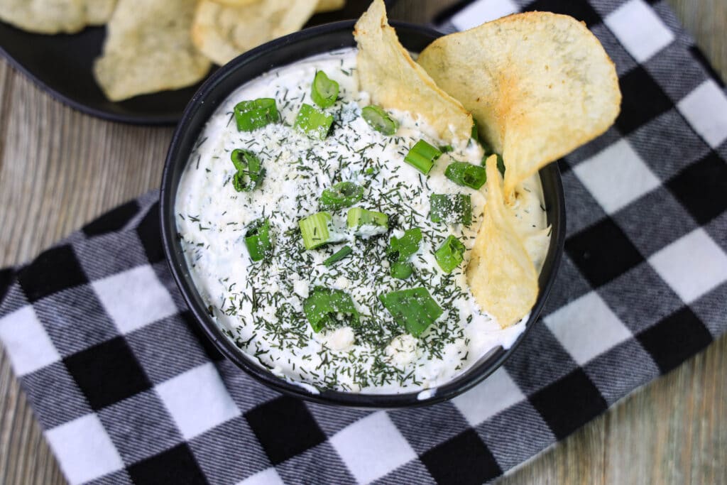 Dill dip in a black bowl on a black and white napkin on a wood table with potato chips 