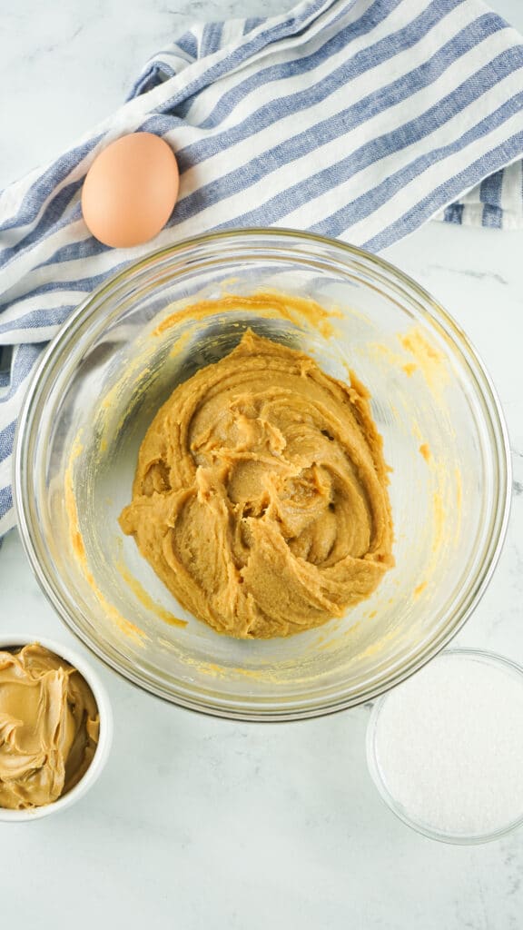 A glass bowl with peanut butter cookie dough mixed up in it. a small cup of peanut butter and a separate cup of sugar and a brown egg are on the table next to the bowl. a blue and white dish towel is under the egg.