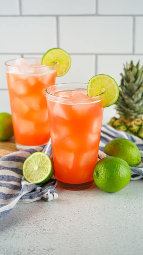 2 pink colored drinks in glasses on a table. The drinks are garnished with lime wedges and have limes on the table around them. 