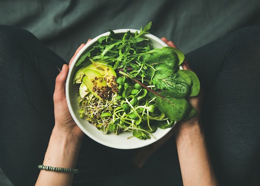 Green vegan breakfast meal in bowl with spinach, arugula, avocado, seeds and sprouts. Girl in leggins holding plate with hands visible, top view. Clean eating, dieting, vegan food concept