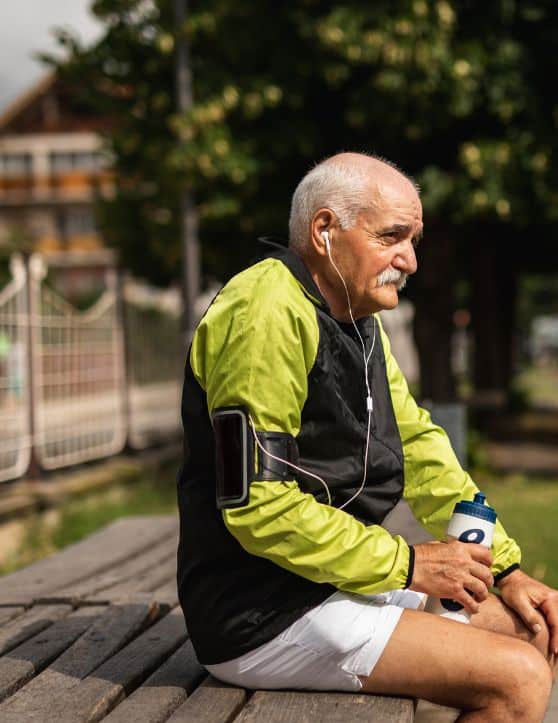 A senior gentleman, donning sportswear, sits comfortably on a wooden bench, engrossed in music playing through his headphones.