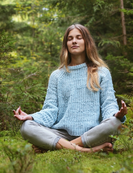 Women sitting in the grass surrounded by treed, legs crossed in the Yoga pose called the lotus